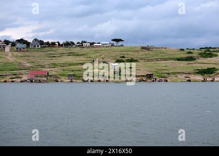 Am ruhigen Ufer des Kazinga-Kanals bereiten die Fischer ihre Boote unter bewölktem Himmel sorgfältig vor, während rustikale Dorfhäuser die üppige Vegetation säumen Stockfoto