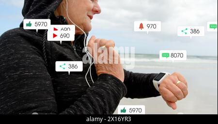 Bild von Benachrichtigungsbalken über einer älteren kaukasischen Frau mit Kopfhörern, die Smartwatch am Strand verwenden Stockfoto