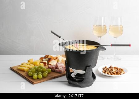 Fonduekanne mit leckerem geschmolzenem Käse, Gabeln, Wein und verschiedenen Snacks auf weißem Holztisch Stockfoto