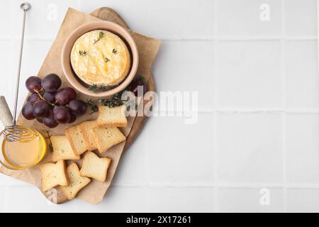 Leckerer gebackener Camembert in der Schüssel, Croutons, Trauben, Honig und Thymian auf weiß gekachelten Tisch, Blick von oben. Leerzeichen für Text Stockfoto