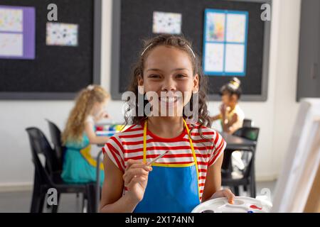 Im Kunstunterricht lächelt ein birassisches junges Mädchen mit einem Pinsel in die Kamera Stockfoto