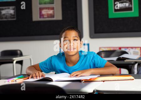 In der Schule saß ein junger männlicher Schüler mit birassischem Hintergrund an einem Schreibtisch in einem Klassenzimmer und lächelte Stockfoto