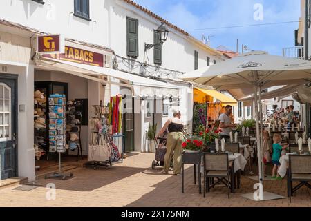 Touristen gehen durch eine enge, gemütliche Straße mit Restaurants und Geschäften im Dorf Fornells auf der spanischen Insel Menorca. Stockfoto