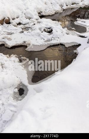 Ein schmaler kleiner Fluss im Winter, bedeckt mit Schnee und Eis, das Eis auf dem Fluss ist mit einem Kunststoff-geogitter verstärkt Stockfoto