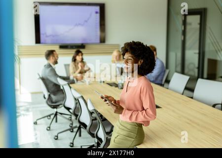 Ccheerful Afroamerikanerin leitet ein Meeting und hält sich an einem digitalen Tablet fest, während Kollegen hinter ihr Gespräche führen. Stockfoto