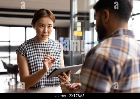 Asiatische Frau mittleren Alters zeigt einem jungen asiatischen Mann in einem modernen Geschäftsbüro ein Tablet Stockfoto
