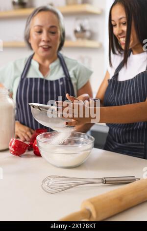 Asiatische Großmutter und birassische Teenager-Enkelin mit Schürzen backen zu Hause zusammen Stockfoto