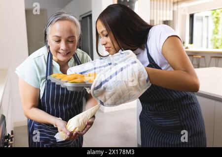 Asiatische Großmutter und birassische Teenager-Enkelin mit Schürzen backen zu Hause zusammen Stockfoto