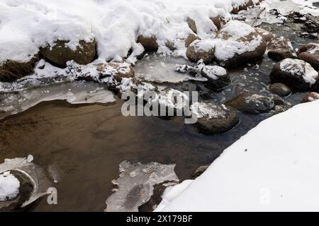 Ein schmaler kleiner Fluss im Winter, bedeckt mit Schnee und Eis, das Eis auf dem Fluss ist mit einem Kunststoff-geogitter verstärkt Stockfoto