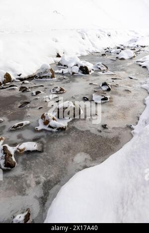 Ein schmaler kleiner Fluss im Winter, bedeckt mit Schnee und Eis, das Eis auf dem Fluss ist mit einem Kunststoff-geogitter verstärkt Stockfoto