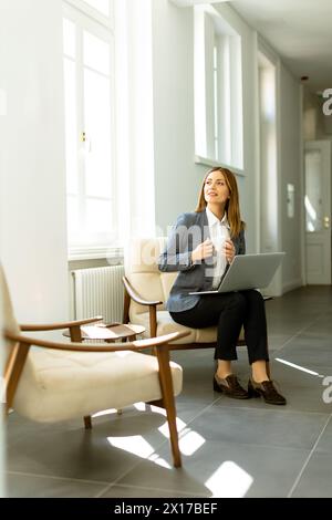 Die professionelle Profi schlürft ihren Kaffee, während sie sich mit ihrem Laptop beschäftigt, während sie sich bei weichem Tageslicht sonnt. Stockfoto