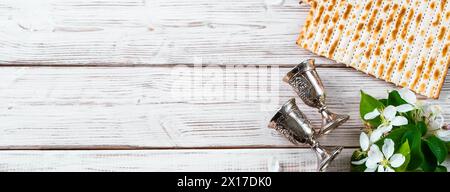 Happy Passover Flatlay. Jüdisches Matzobrot und Weingläser auf weißem hölzernem Hintergrund. Weihnachtsgrußkarte mit Blumen. Kopierbereich, Bannerformat. Stockfoto