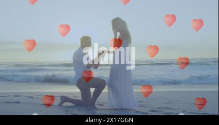 Bild von Ballons aus rotem Herzen, die über ein Paar im Liebesmann schweben und eine Frau mit einem Ring am Strand vorschlagen Stockfoto