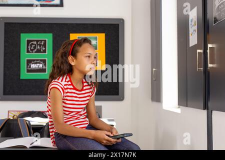 In der Schule, im Klassenzimmer, sieht ein junges birassisches Mädchen, das ein Smartphone hält, nachdenklich aus Stockfoto