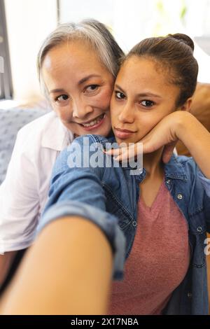 Asiatische Großmutter, die zu Hause eine birassische Teenager-Enkelin umschließt, beide lächeln vor der Kamera Stockfoto