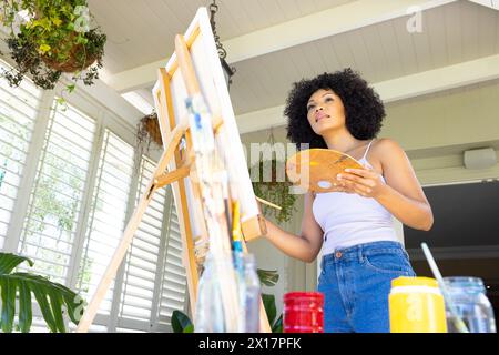 Junge birassische Frau, die zu Hause auf Leinwand malt und Palette hält Stockfoto