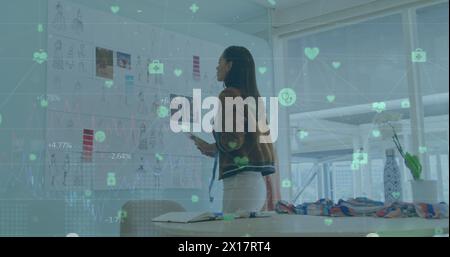 Bild von verbundenen Ikonen und Zahlen über birassische Frau, die das Design des Kleides im Büro untersucht Stockfoto