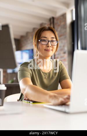 Asiatische Frau mittleren Alters mit Brille, die in einem modernen Geschäftsbüro an einem Laptop arbeitet Stockfoto
