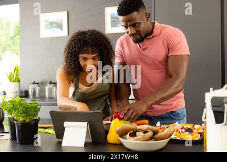 Diverse Paare bereiten in der Küche zu Hause Essen zu und suchen nach Rezepten auf Tablets Stockfoto