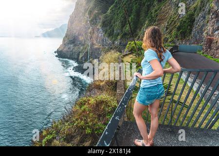 Beschreibung: Sportliche Frau mit Blick auf den Wasserfall, der in atmosphärischer Morgenatmosphäre ins Meer fließt. Aussichtspunkt Véu da Noiva, Insel Madeira, Portuga Stockfoto