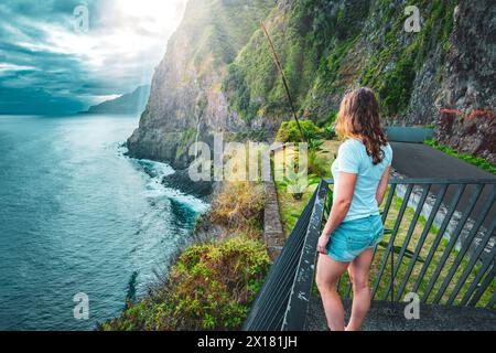 Beschreibung: Sportliche Frau mit Blick auf den Wasserfall, der in atmosphärischer Morgenatmosphäre ins Meer fließt. Aussichtspunkt Véu da Noiva, Insel Madeira, Portuga Stockfoto