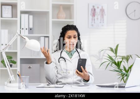 Eine Ärztin in einem weißen Labormantel zeigt einen verwirrten Ausdruck, während sie ihr Smartphone in einem modernen Klinikbüro ansieht. Pflanzen und Anatomiediagramme im Hintergrund. Stockfoto