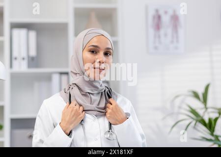 Ein Porträt einer widerstandsfähigen und selbstbewussten muslimischen Ärztin, die in einem gut ausgestatteten medizinischen Büro Hijab trägt und Professionalität und Pflege ausstrahlt. Stockfoto