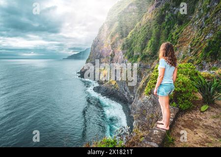 Beschreibung: Sportliche Frau mit Blick auf den Wasserfall, der in atmosphärischer Morgenatmosphäre ins Meer fließt. Aussichtspunkt Véu da Noiva, Insel Madeira, Portuga Stockfoto