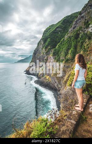 Beschreibung: Sportliche Frau mit Blick auf den Wasserfall, der in atmosphärischer Morgenatmosphäre ins Meer fließt. Aussichtspunkt Véu da Noiva, Insel Madeira, Portuga Stockfoto