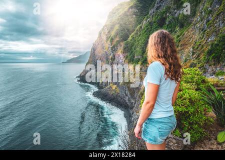 Beschreibung: Sportliche Frau mit Blick auf den Wasserfall, der in atmosphärischer Morgenatmosphäre ins Meer fließt. Aussichtspunkt Véu da Noiva, Insel Madeira, Portuga Stockfoto