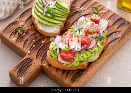 Avocado toast auf einem Holzbrett mit Frischkäse, Radieschen und Feta, garniert mit roten Zwiebeln und frisch gemahlenem Pfeffer Stockfoto
