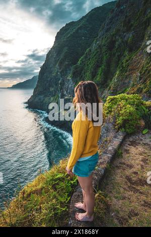 Beschreibung: Sportliche Frau mit Blick auf den Wasserfall, der in atmosphärischer Morgenatmosphäre ins Meer fließt. Aussichtspunkt Véu da Noiva, Insel Madeira, Portuga Stockfoto