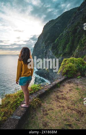 Beschreibung: Sportliche Frau mit Blick auf den Wasserfall, der in atmosphärischer Morgenatmosphäre ins Meer fließt. Aussichtspunkt Véu da Noiva, Insel Madeira, Portuga Stockfoto