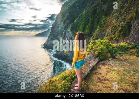 Beschreibung: Sportliche Frau mit Blick auf den Wasserfall, der in atmosphärischer Morgenatmosphäre ins Meer fließt. Aussichtspunkt Véu da Noiva, Insel Madeira, Portuga Stockfoto