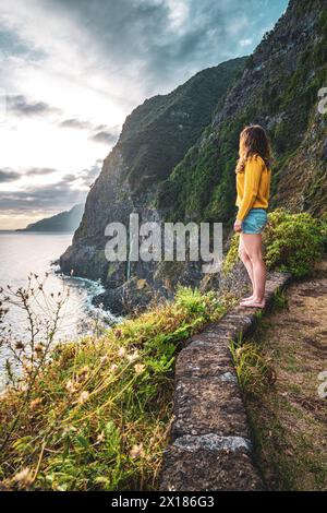 Beschreibung: Sportliche Frau mit Blick auf den Wasserfall, der in atmosphärischer Morgenatmosphäre ins Meer fließt. Aussichtspunkt Véu da Noiva, Insel Madeira, Portuga Stockfoto