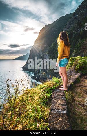 Beschreibung: Sportliche Frau mit Blick auf den Wasserfall, der in atmosphärischer Morgenatmosphäre ins Meer fließt. Aussichtspunkt Véu da Noiva, Insel Madeira, Portuga Stockfoto