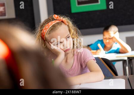 In der Schule sieht das junge weiße Mädchen mit lockigen blonden Haaren im Klassenzimmer nachdenklich aus Stockfoto