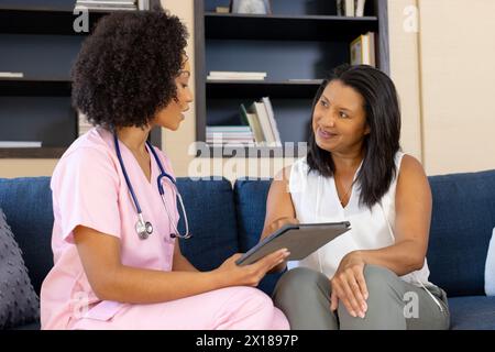 Die birassische Krankenschwester, die eine Tablette hält, spricht mit reifer birassischer Frau zu Hause Stockfoto