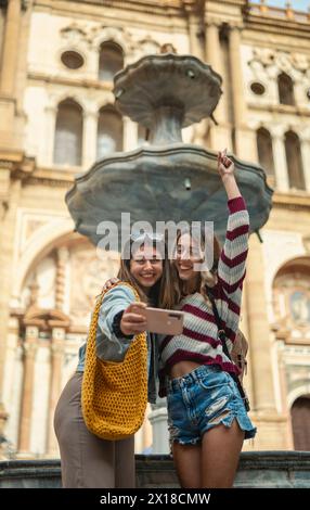 Zwei kaukasische Touristenmädchen, die einen lächelnden Arm heben und ein Selfie vor einem Brunnen in der Kathedrale von Malaga machen. Stockfoto