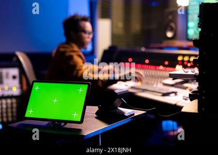 afroamerikanischer Tontechniker, der Musik neben dem Greenscreen-Display auf Gadget bearbeitet und über die Mischkonsole im Kontrollraum bedient. Junger Produzent, der im Studio Knöpfe und Schieberegler drückt. Stockfoto