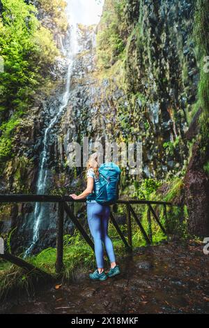 Beschreibung: Sportlicher Tourist mit Rucksack lehnt sich an den hölzernen Zaun des Aussichtspunkts und beobachtet den grün bewachsenen Risco-Wasserfall. 25 Fontes Wasserfälle, Stockfoto