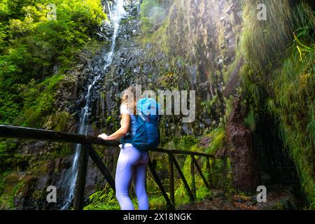 Beschreibung: Frau Touristin mit Rucksack lehnt sich an den hölzernen Zaun des Aussichtspunkts und beobachtet den grünen, bewachsenen Risco-Wasserfall. 25 Fontes Wasserfälle, Ma Stockfoto