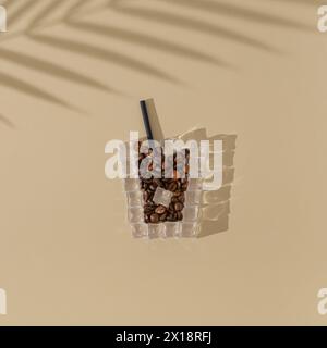 Eine Tasse Eiskaffee aus Eiswürfeln und Kaffeebohnen mit Palmenblattschatten auf sandfarbenem Hintergrund. Kreatives Konzept für den Sommer Minimal. Stockfoto
