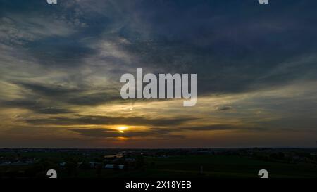 Dramatischer Sonnenuntergang, der Ein warmes Leuchten über Einer ländlichen Landschaft mit Silhouetten von Bäumen und Gebäuden unter Einem wolkenbedeckten Himmel zaubert. Stockfoto