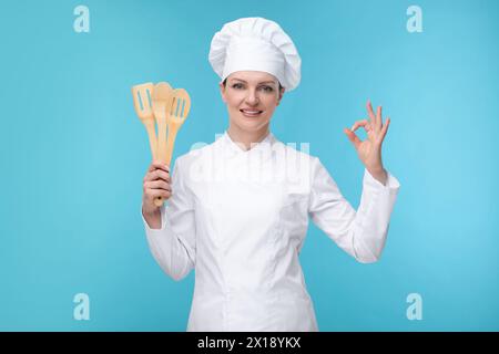 Glücklicher Koch in Uniform, der Holzutensilien auf hellblauem Hintergrund hält Stockfoto