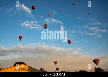 Eine Flotte lebendiger Heißluftballons steigt bei Sonnenuntergang über einem lokalen Bauernmarkt in den Himmel und verleiht dem Tag einen Hauch Magie Stockfoto