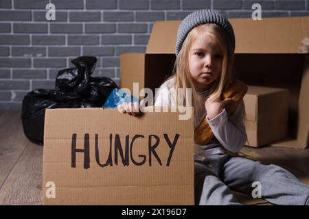 Obdachloses kleines Mädchen mit Brot, das um Essen bettelt, während es auf dem Boden sitzt Stockfoto