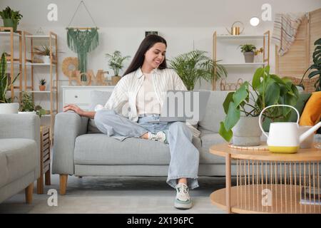 Junge Frau, die auf dem Sofa mit Laptop in der Nähe des Tisches mit der Gießkanne im Wohnzimmer sitzt Stockfoto