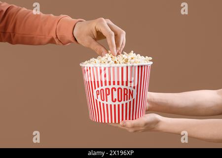 Männliche Hand, die Popcorn aus dem Eimer nimmt weibliche Hände auf braunem Hintergrund Stockfoto