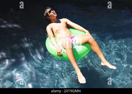 Ein asiatischer Teenager, der eine Sonnenbrille trägt und sich zu Hause auf einem grünen Schwimmer in einem Pool entspannt Stockfoto